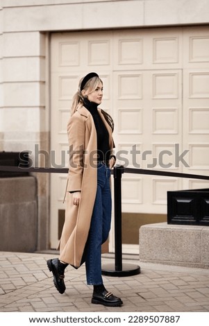 Full length portrait of young beautiful woman walking outdoors stylishly dressed in spring or autumn clothes cashmere beige coat, jeans, loafers, turtleneck. Female street style fashion Royalty-Free Stock Photo #2289507887