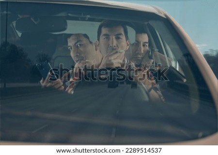 A Latin male driver is shown driving dangerously while smoking and using his phone, endangering himself and others on the road. This cautionary image highlights the risks of distracted driving. Royalty-Free Stock Photo #2289514537