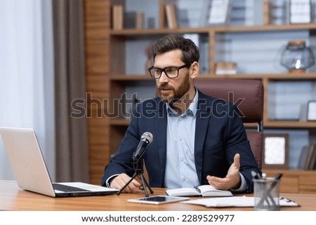 Mature businessman inside office with professional microphone and laptop recording audio podcast and training seminar, experienced boss financier sitting at desk in office video call online stream.