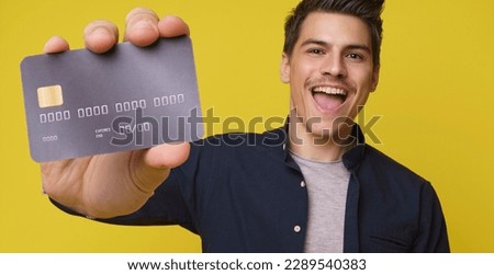 Impressed handsome young person holds in hand debit credit card happy open mouth isolated on yellow color background. Close up of big bank card in hand of 20s young man hipster in blue shirt Royalty-Free Stock Photo #2289540383