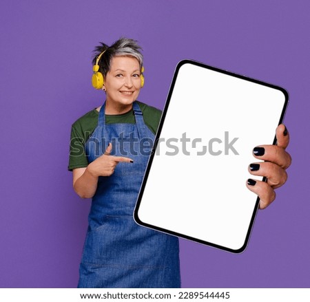 50s mature smiling business woman in jeans apron holding big digital tablet computer showing blank empty mock up screen display for advertising or web service standing isolated on violet background.