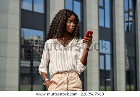 Happy young African model using smartphone standing outdoors. Pretty black business woman holding cell phone doing online shopping, checking apps looking at cellphone technology device in city.