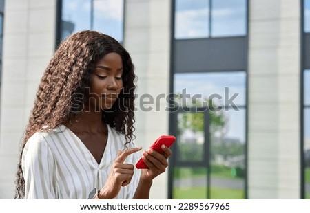 Young African lady using smartphone standing outdoors. Pretty black business woman holding cell phone doing online shopping, scrolling, browsing looking at cellphone tech device in city.