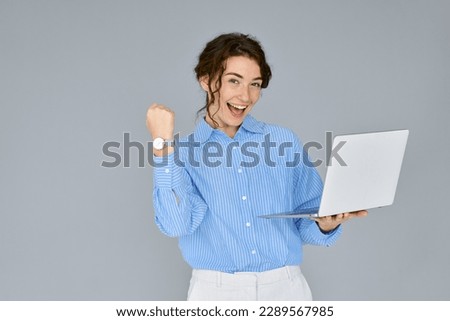 Young happy excited business woman executive holding laptop computer raising fist in yes gesture celebrating online win, business success, good result or reward standing isolated on grey background.