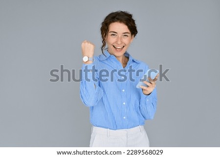 Young happy confident professional business woman executive feeling excited winner looking at cellphone using mobile phone receiving new job offer, celebrating online money win standing at gray wall.