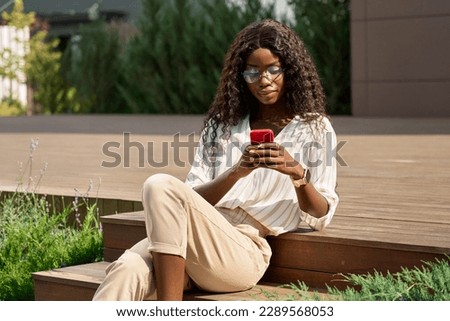 Young African black happy woman model holding cell phone using mobile apps for ecommerce shopping in online applications, communicating online on cellphone tech device sitting outside in city park.