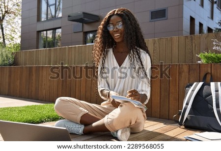 Happy black young woman model, pretty curly African female university student elearning using laptop computer studying outside campus on sunny day, remote learning online, having hybrid call.