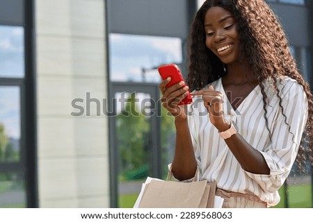 Happy young black African woman model standing on city street holding shopping bags using cell phone mobile app buying ordering fashion clothes online in ecommerce digital store on smartphone.