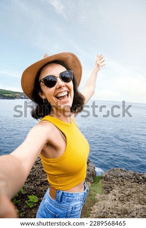 Joyful and smiling young woman traveler taking selfie with phone during summer vacation in seaside location. Vertical.
