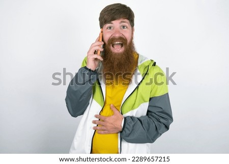 Smiling red haired man wearing sportswear standing over white studio background talks via cellphone, enjoys pleasant great conversation. People, technology, communication concept