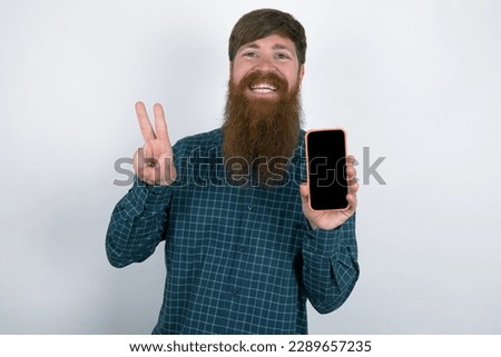 red haired man wearing plaid shirt over white studio background holding modern device showing v-sign