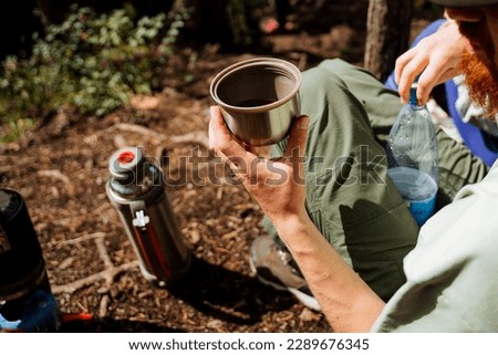 Hand holding a glass of cold water, a thermo tea mug, a thermos of tea, breakfast in nature, hiking alone, camping in the forest, drinking tea in nature. High quality photo