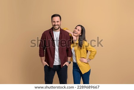 Cheerful young girlfriend with hand on boyfriend's shoulder laughing and looking at camera. Happy young couple posing and standing ecstatically against beige background Royalty-Free Stock Photo #2289689667