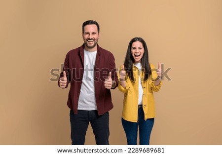 Charming young woman and man dressed in casuals showing thumbs up and laughing while standing over beige background. Cheerful couple looking at camera and showing approval gesture. Thumbs up!
