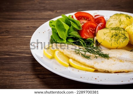 Fish dish - fried cod with boiled potatoes and fresh vegetables on wooden table 