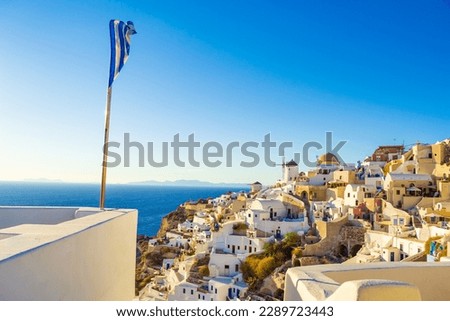 Greek flag over beautiful Oia village architecture,Santorini.It has become globally famous for its authentic Cycladic beauty, whitewashed houses, labyrinth-like alleys, and amazing hotels.