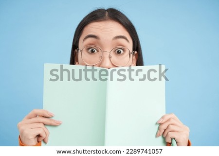 Cute asian woman in glasses, hides her face behind notebook and smiles with eyes, curious gaze, stands over blue background. Students and education concept. Royalty-Free Stock Photo #2289741057