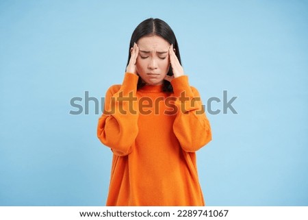 Frustrated asian woman holds hands on head, has headache, looks miserable, cant handle pressure, feels distressed, stands over blue background. Mental health concept. Royalty-Free Stock Photo #2289741067