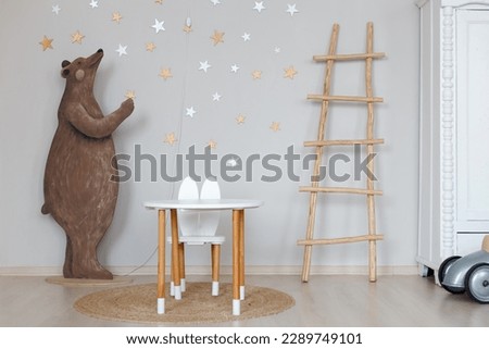 Children's room interior with decor. The white wall is decorated with stars and a picture of a bear. There is a children's table and chair on the floor. A wooden ladder.