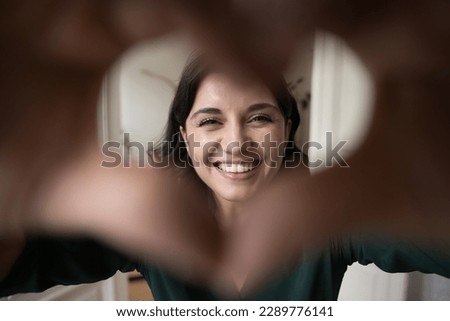 Close up face Armenian overjoyed woman show symbol of love with joined fingers, sign of happiness and romance, look through heart shaped frame, charity, volunteering, professional medical services ad
