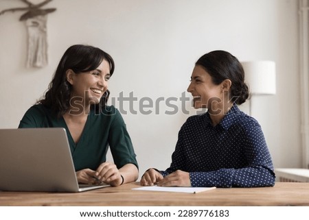 Happy multiethnic women have fun seated at desk in workplace. Female teammates, distracted from work, tell jokes, laughing, having goods friendly work relations, engaged in teamwork and communication Royalty-Free Stock Photo #2289776183
