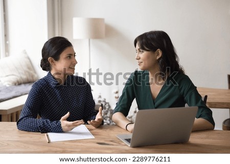 Serious Indian mentor or teacher talk to female colleague teach intern, discussing new project or task sit at desk in office. Two diverse coworkers working together, cooperate, engaged in team-work Royalty-Free Stock Photo #2289776211