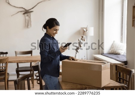 Millennial Indian woman sending cardboard box, prepare goods for shipment using smartphone, calling for courier to pick up parcel, use on-line mobile application delivery electronic services. Dispatch Royalty-Free Stock Photo #2289776235