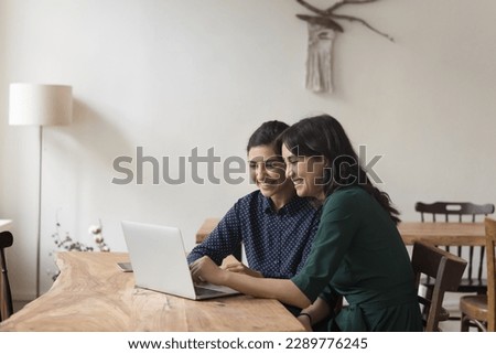 Two pretty multiethnic women colleagues sit at desk watch online presentation on laptop, share ideas enjoy amusing on-line content during break at workplace. Workflow using modern tech, fun, teamwork Royalty-Free Stock Photo #2289776245