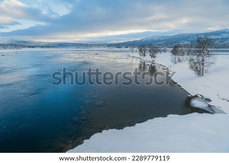 Winter landscape in Tjåmotis with mountains in background, sun shining over the mountains, creek with open water, sky with nice colors reflecting in the water, Swedish Lapland, Sweden Royalty-Free Stock Photo #2289779119