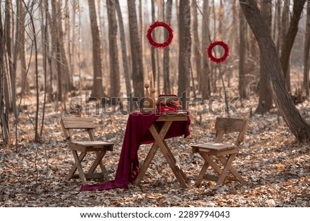 autumn pre-wedding decoration for photo shoot. wooden chair and table with red tablecloth with red circle boho accessories hanging from the tree. Yalda decoration outdoor.