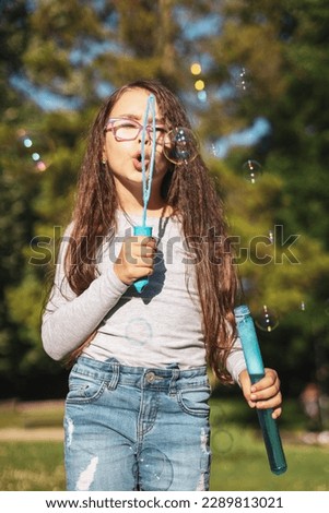 Portrait of a beautiful caucasian girl blowing soap bubbles with colored bokeh while standing in the park on the playground against the background of blurry trees, close-up side view with selective fo