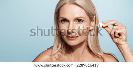 Portrait of elrderly woman takes care of her skin with cosmetic toner, hyaluronic acid aha serum for skin lifting and anti-aging effect, holds dropper near face, blue background. Royalty-Free Stock Photo #2289816419