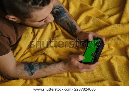 Young man lies inside on yellow blanket on bed and plays using phone and joystick, top view. Hands with multicolored tattoos close-up. Guy Uses Green Mock-up Screen Smartphone.