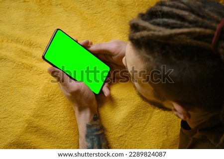 Guy Uses Green Screen Smartphone. Young man lies on yellow blanket and uses phone to work or browse the Internet online, watching movie or TV series, top view. Copy space for advertising and design.