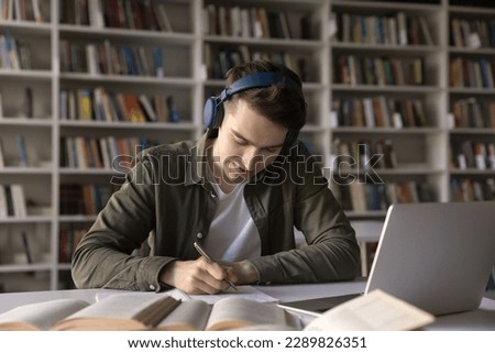 Focused student guy sit at desk work hard on research project in library, listen audio lesson through headphones improve language skills, get ready for admission, gain new knowledge use modern tech Royalty-Free Stock Photo #2289826351