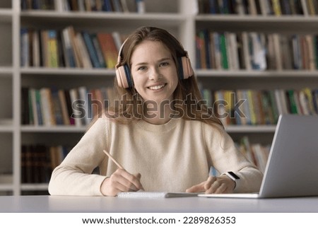 Pretty teen 16s girl prepare for university exams or college admission use modern tech, sit at desk in library wear headphones look at camera. Portrait of excellent high school student, self-education Royalty-Free Stock Photo #2289826353