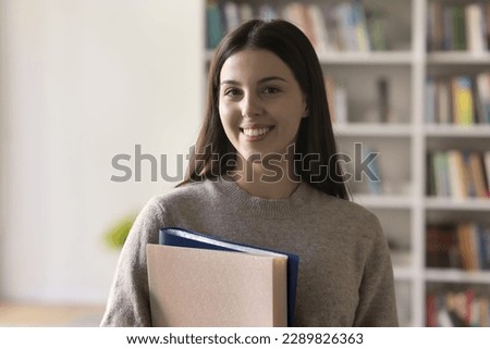 Head shot portrait of pretty teenager, student girl holding folders pose standing in campus library smile looking at camera. High school or university education, professional knowledge, studentship