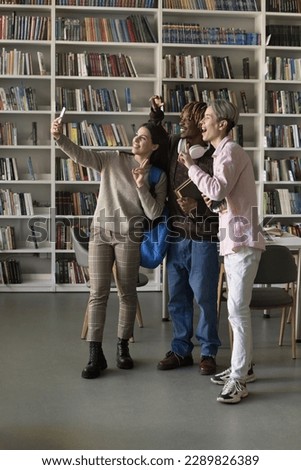 Three multi ethnic friends, girl and guys students standing in library staring at cellphone screen make selfie pictures, gesturing, record video for channel. Generation Z and modern tech usage, fun