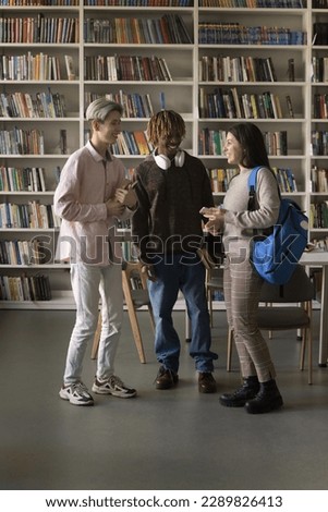 Three multi racial teenage, diverse schoolmates talking standing in college or public library, share news, discuss joint project or schedule, having good friendly relations. Education, communication Royalty-Free Stock Photo #2289826413