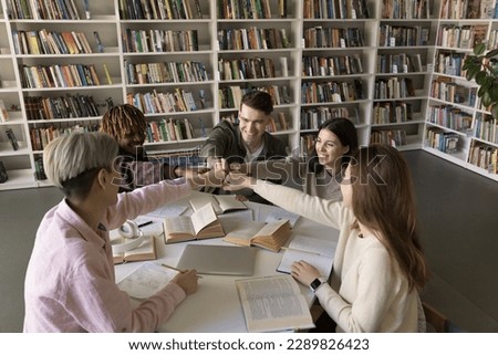 Happy multi ethnic group of university students sit at desk with books in library, showing team unity, do joint project, smile, work on assignment, celebrating success, hands fist bump join together Royalty-Free Stock Photo #2289826423