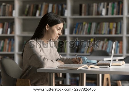 Girl prepares for exams in university library, writing exercise, makes notes, jotting information learning theory, sit at desk looks at laptop, reading text, do assignment. New knowledge, modern tech Royalty-Free Stock Photo #2289826455