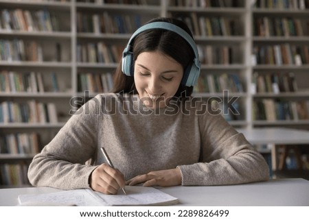 Smiling attractive student girl listen audio task, improve foreign language knowledge sit at desk writes exercise, take notes, studying, e-learn in library. Modern tech usage for new skills gaining Royalty-Free Stock Photo #2289826499