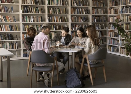 In cozy college library five multi ethnic students, girls and guys engaged in homework making, preparing for exams seated at desk on bookshelves background. Get higher education, studentship, teamwork