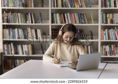 Smart student girl wear wireless headphones work on graduation project, creative essay in modern library, listen audio lesson, writes notes, preparing for admission to higher institution. Education Royalty-Free Stock Photo #2289826519