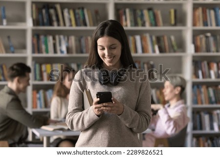 Teenage student girl standing in library or classroom use smart phone, search information, take break, read sms from friend, chatting on-line, check timetable through mobile apps. Modern tech, studies Royalty-Free Stock Photo #2289826521