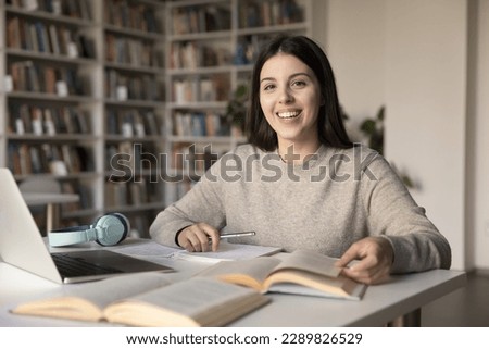 Portrait of pretty teen girl student studying in library, doing homework sit at desk smile staring at camera enjoy studies, gains new knowledge, practicing language, prepare for university admission Royalty-Free Stock Photo #2289826529