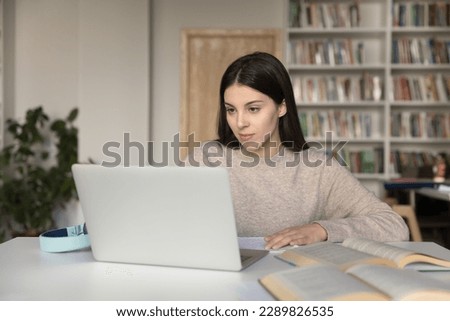 Attractive student girl sits at desk in library prepare for university exams or high school admission using laptop looks focused, makes exercise, browse information, do task. E-learning, modern tech Royalty-Free Stock Photo #2289826535