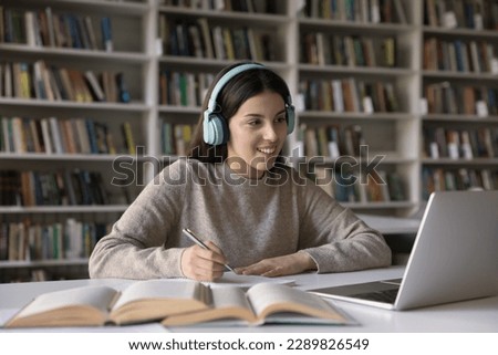 Pretty student teen 17s girl wear headphones e-learns subject, prepare for exams seated at desk studying through videoconference with tutor using laptop in library. Gen Z engaged in on-line education Royalty-Free Stock Photo #2289826549