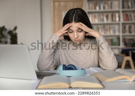 Schoolgirl sit at desk with textbook in library cramming, prepare for exams, think, solve math task, read theory, holds her head feels overworked, tired from studying or cramming. Information overload
