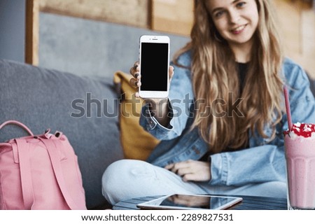 Portrait of woman in cafe drinking smoothie and showing mobile phone, focus on smartphone screen. Girl showing application. Royalty-Free Stock Photo #2289827531